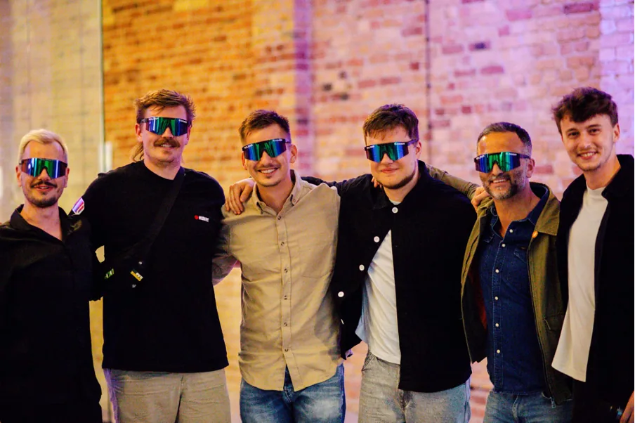 Six men in matching mirrored sport sunglasses posing arm in arm in front of a brick wall