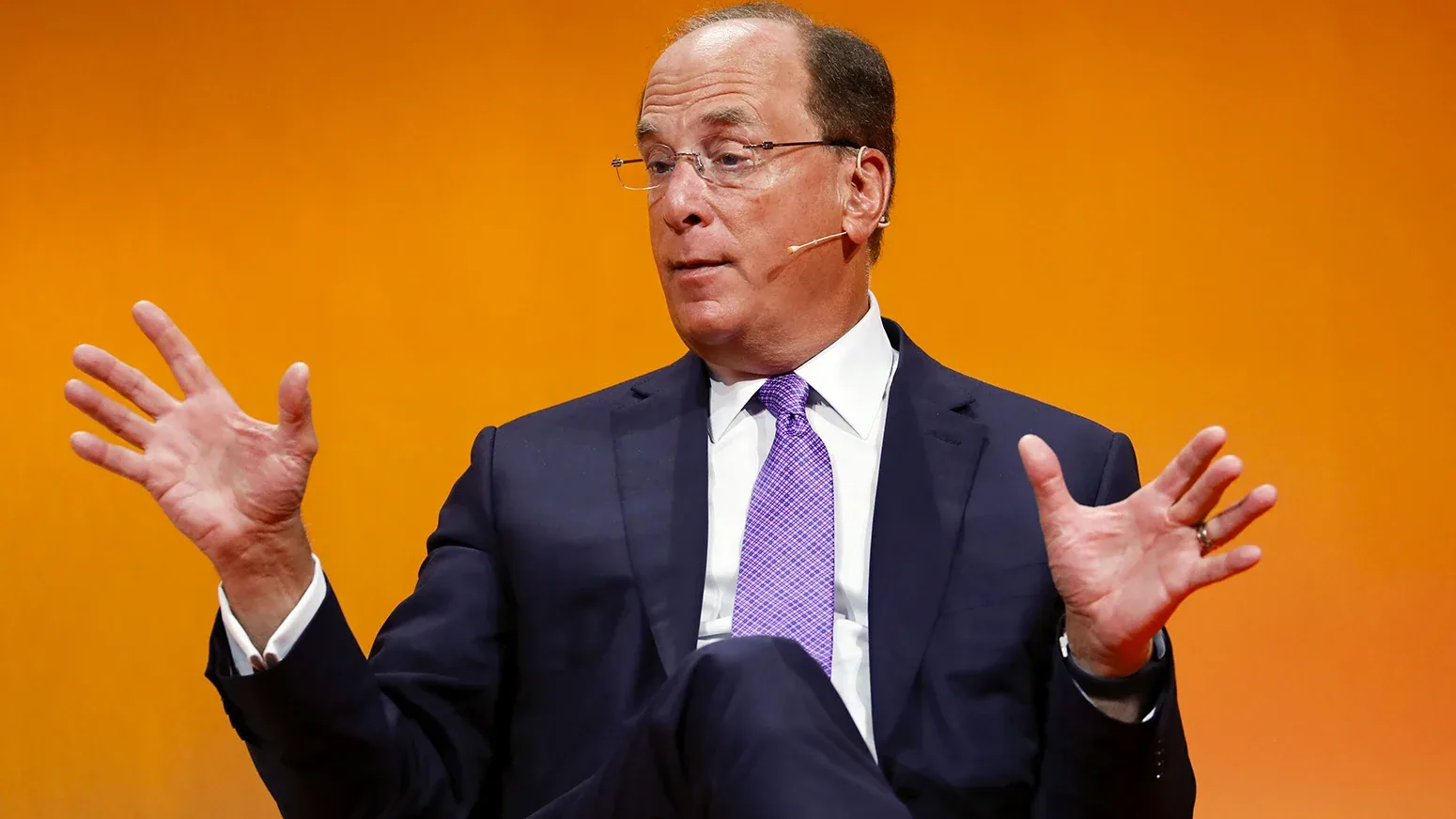 Man in a dark suit and purple tie gesturing as he speaks on a stage with an orange backdrop